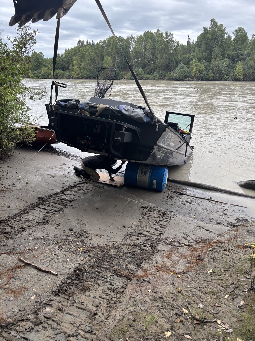 Chad lying under a boat hoisted by excavator straps, applying sealant to a hole in the hull
