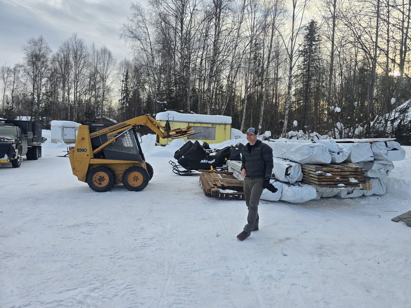 Chad standing beside stacked lumber while a Case 1840 skid steer loads material behind him