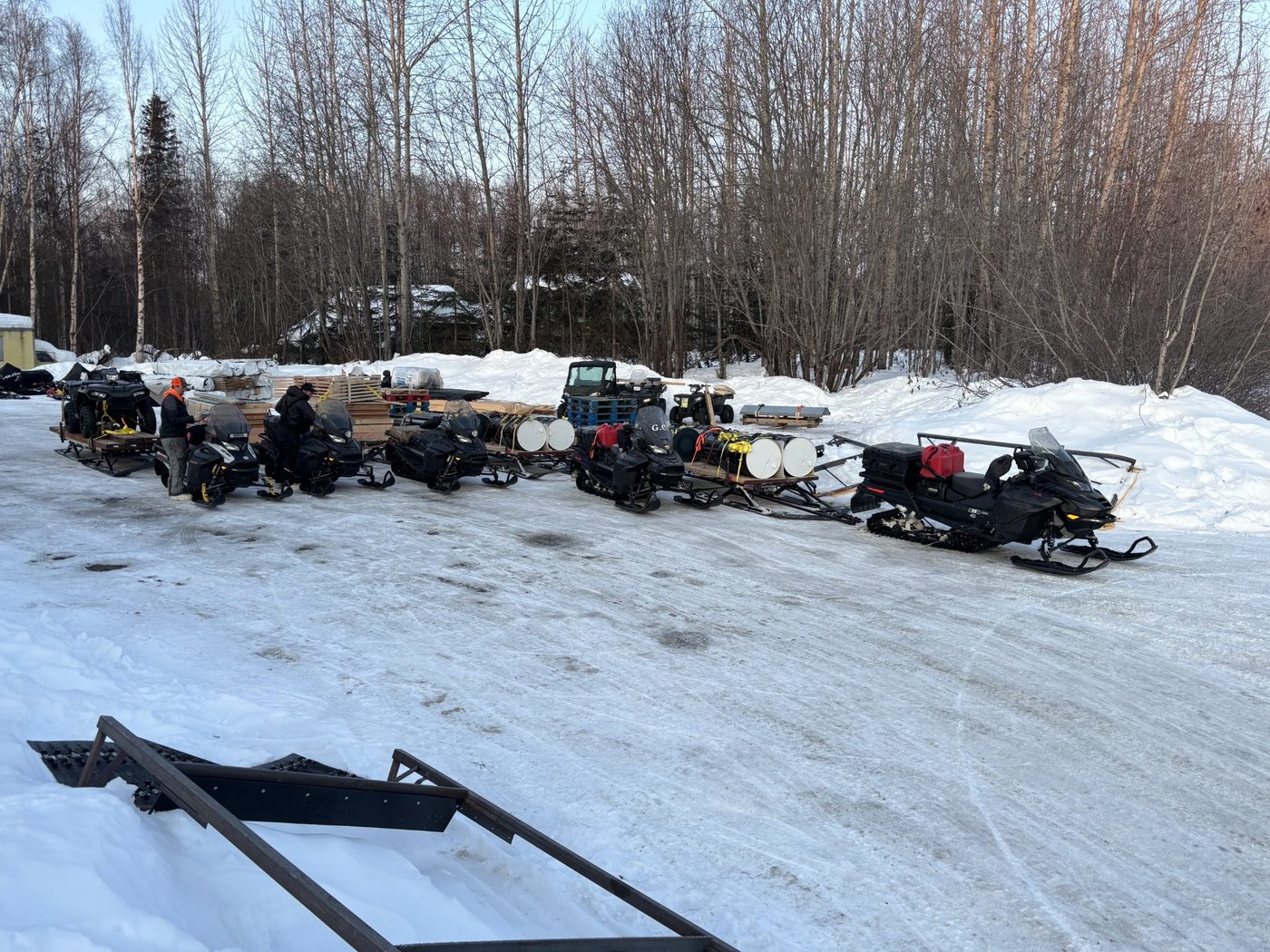 A line of snow machines hitched to freight sleds loaded with barrels and lumber