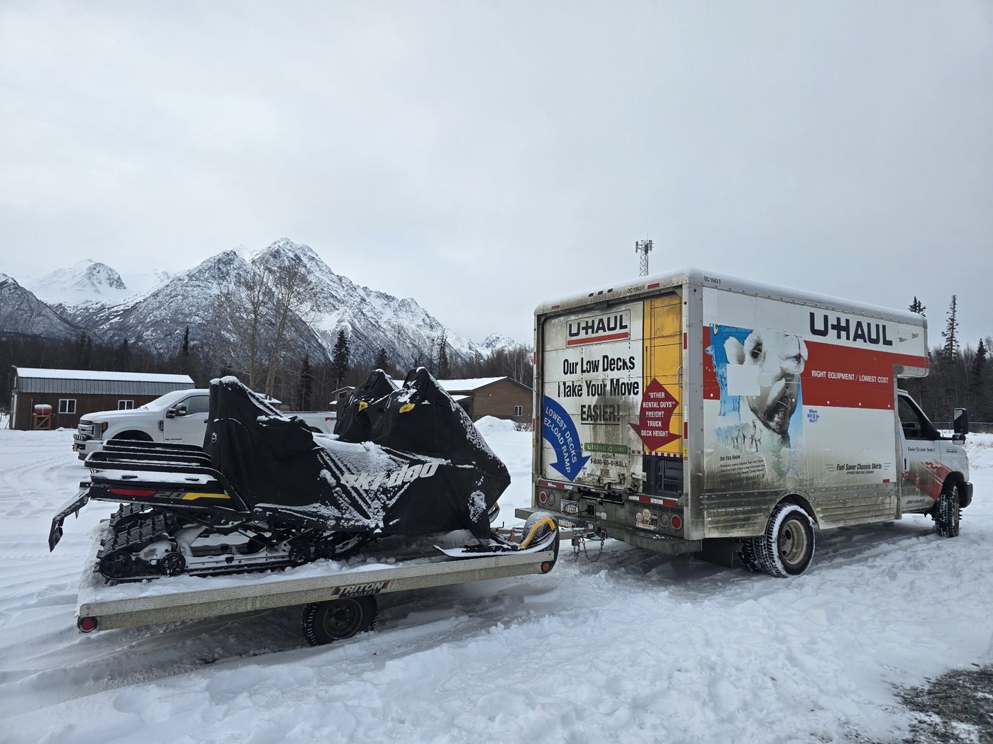 U-Haul truck parked next to a trailer loaded with snow machines in a snowy yard